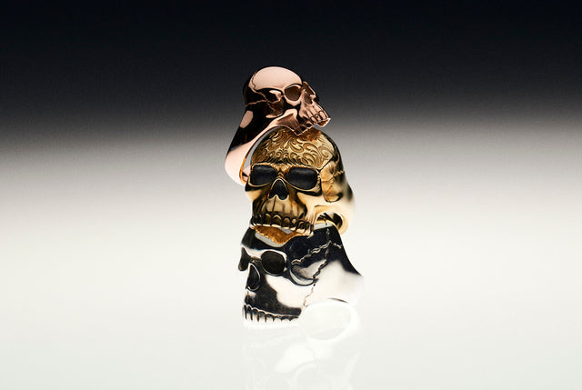A skull ring in rose gold, a yellow gold skull ring with a tattoo design engraved on it's head and a silver large skull ring. They are placed one on top of the other.